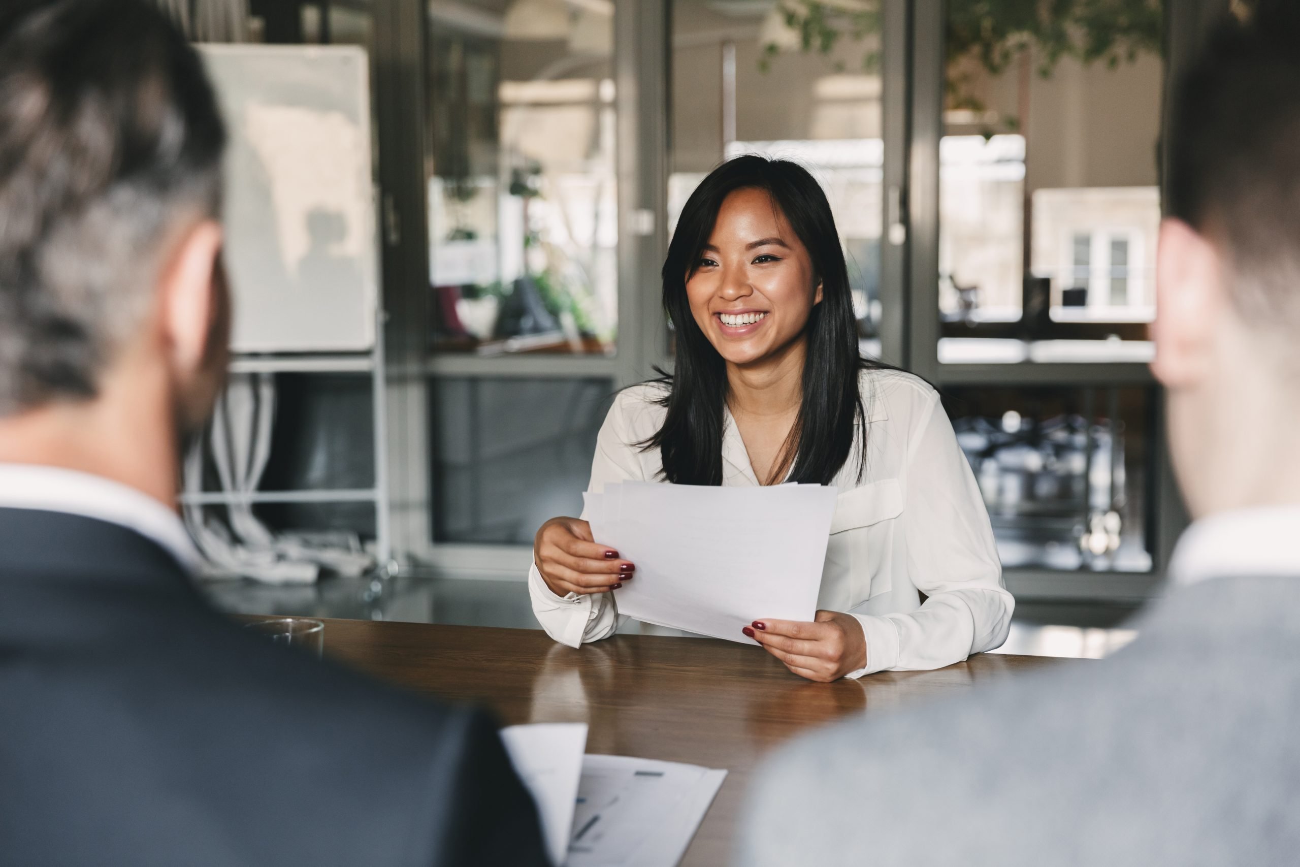 Young woman smiling and holding resume, while sitting in front of directors during job interview