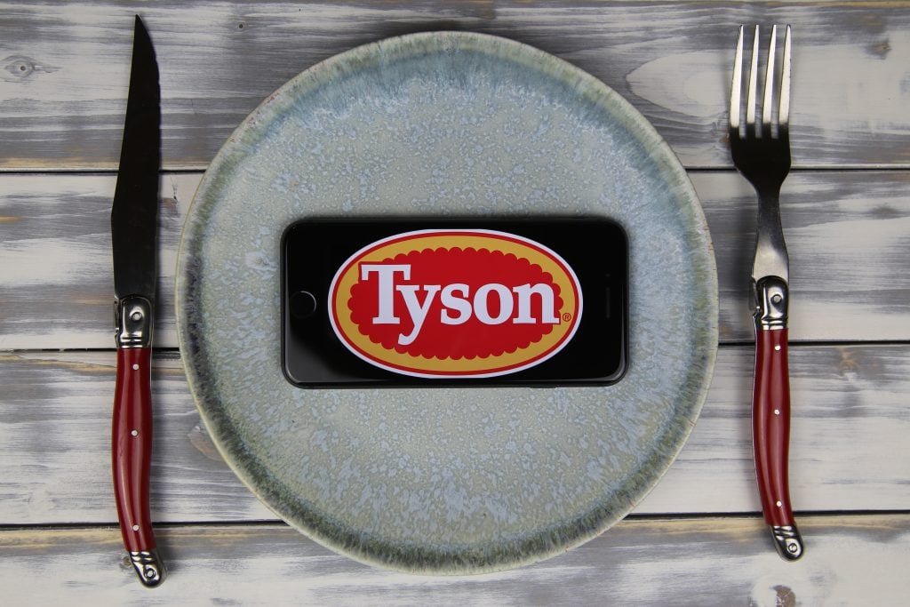  Closeup of mobile phone on plate and wood table, cutlery with logo lettering of Tyson foods company (focus on center)