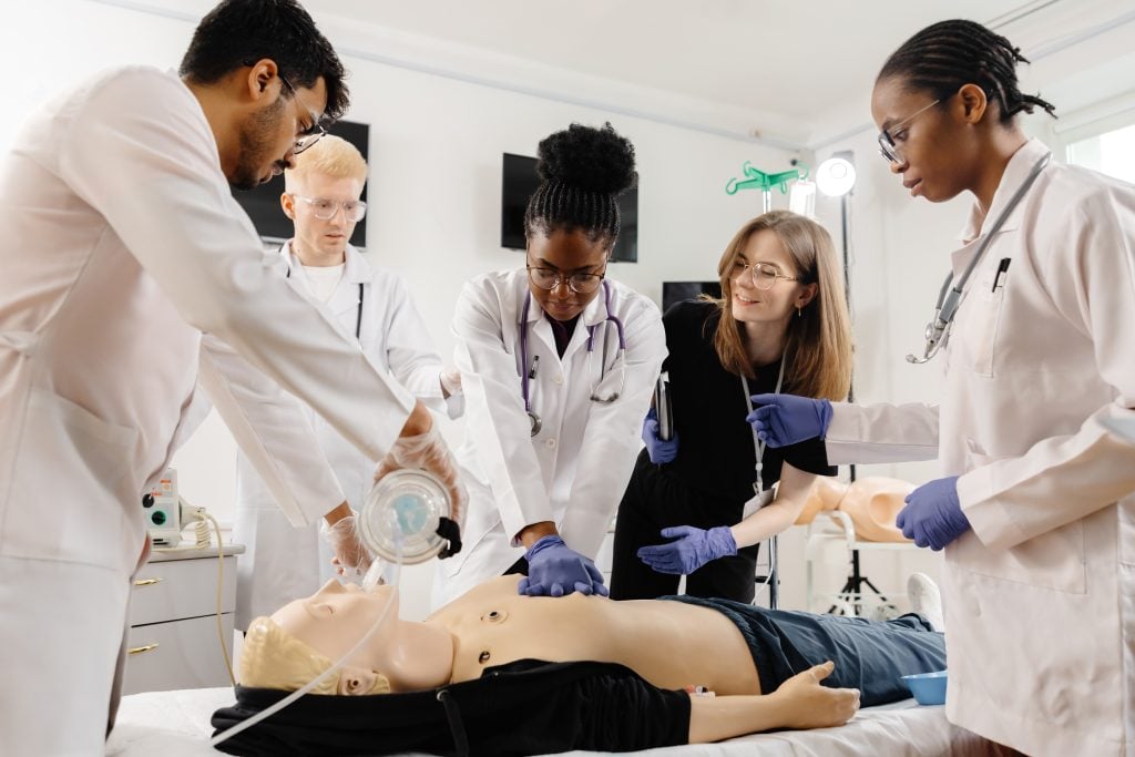 A group of medical personnel is gathered around a CPR training dummy, practicing life-saving techniques in a medical simulation setting.